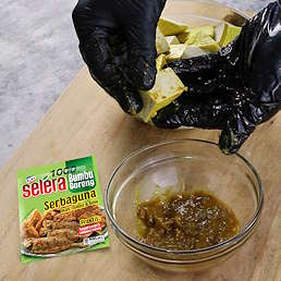 Tangan bersarung membumbui potongan tahu dengan Selera Bumbu Goreng Serbaguna dari Kobe, terlihat mangkuk bumbu dan kemasan produk