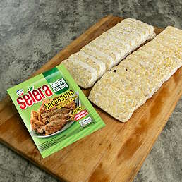 Potongan tempe di atas talenan kayu dan kemasan Selera Bumbu Goreng Serbaguna dari Kobe ukuran 70g