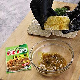 Tangan bersarung mengoles bumbu pada potongan tempe, terlihat mangkuk bumbu dan kemasan Selera Bumbu Goreng Serbaguna dari Kobe